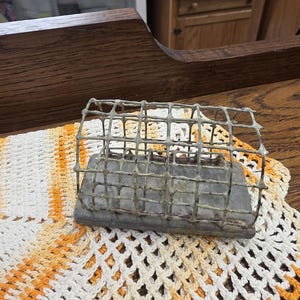 Op de afbeelding: Een metalen kooi-achtig object met een rechthoekige basis, mogelijk een kleine diervoederbak of decoratief stuk. De kooi is gemaakt van gaas en staat op een lichtgekleurd, gehaakt kleedje met oranje en gele accenten. De achtergrond is een houten oppervlak.