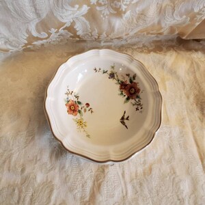May include: A white ceramic bowl with a floral design. The bowl has a scalloped edge and a gold trim. The flowers are in shades of pink, orange, and yellow.