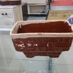 May include: A brown ceramic planter with a white rim and a brick pattern. The planter is rectangular with a slightly rounded bottom.