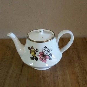 Puede incluir: Tetera blanca con un diseño floral con flores rosas, amarillas y blancas. La tetera tiene un borde dorado alrededor de la tapa y el pico.