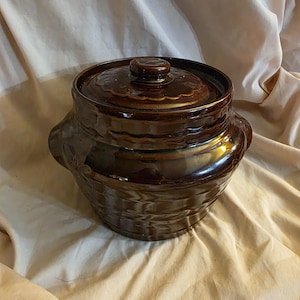 Op de afbeelding: Een bruine keramische pot met een deksel. De pot heeft een getextureerd, golvend ontwerp en een glanzende afwerking.