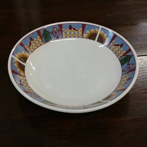 May include: A white ceramic plate with a decorative border featuring sunflowers, birdhouses, and a blue sky design. The plate is oval-shaped and sits on a dark wooden surface.