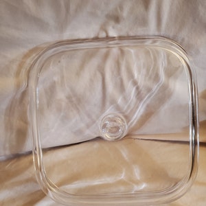 May include: A clear, square glass lid with rounded corners and a small, circular handle. The lid is designed to fit a square dish. The glass is transparent, allowing visibility of the contents below. The lid is likely for a baking dish or casserole.