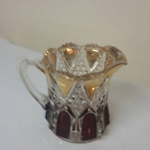 Puede incluir: Una jarra de crema de vidrio transparente con un diseño dorado y rojo. La jarra tiene un borde festoneado y un asa.