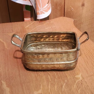 Könnte beinhalten: Ein kleiner, messingfarbener Pflanztopf mit zwei Griffen. Der Topf hat eine gehämmerte Oberfläche und eine leicht abgerundete rechteckige Form.