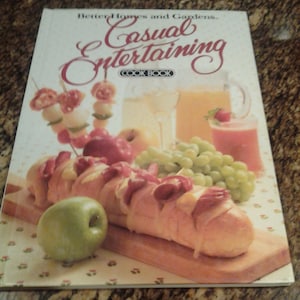 Puede incluir: Un libro de cocina titulado "Better Homes and Gardens Casual Entertaining Cookbook" con una foto de una baguette con queso y carne en una tabla de cortar de madera, rodeada de uvas, manzanas y un vaso de jugo.