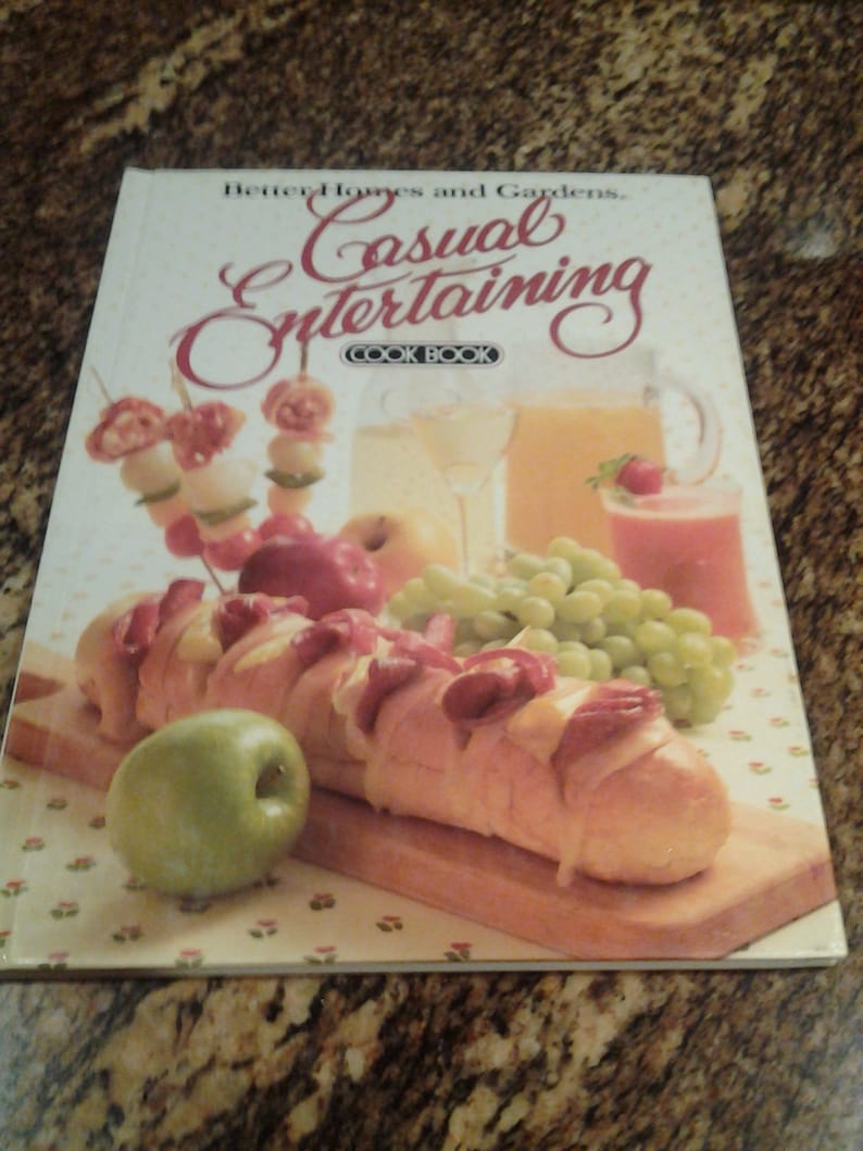 Puede incluir: Un libro de cocina titulado "Better Homes and Gardens Casual Entertaining Cookbook" con una foto de una baguette con queso y carne en una tabla de cortar de madera, rodeada de uvas, manzanas y un vaso de jugo.