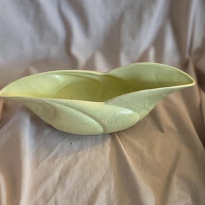 May include: A pale yellow ceramic planter with a speckled finish. The planter is shaped like a leaf and has a wide, shallow bowl.