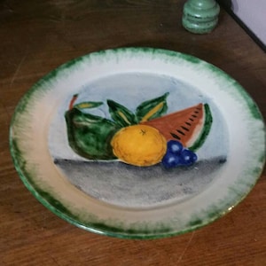May include: A white ceramic plate with a green rim and a painted design of fruit. The design includes a watermelon, a pear, a lemon, and grapes.