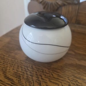 May include: A small, white ceramic jar with a black lid. The jar has a spherical shape and is decorated with thin, curved black lines. The jar is sitting on a wooden surface.