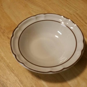 Puede incluir: Un cuenco de cerámica blanco con borde festoneado y borde marrón. El cuenco está vacío y se encuentra sobre una superficie de madera. El diseño es sencillo y clásico, adecuado para servir alimentos.