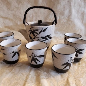 Puede incluir: Un juego de té blanco y negro con una tetera y seis tazas. La tetera tiene un asa negra y una tapa negra. Las tazas tienen un borde negro y un diseño negro de hojas de bambú. El juego está sobre un mantel blanco.