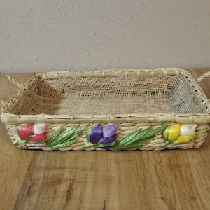 May include: A woven wicker basket with a mesh liner and a decorative border of colorful flowers. The basket has two handles.