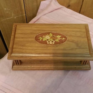 Könnte beinhalten: Eine Holzkiste mit einem floralen Intarsienmuster auf dem Deckel. Die Kiste ist rechteckig und aus hellbraunem Holz gefertigt.