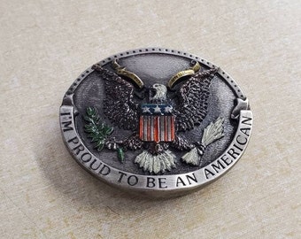 May include: A silver belt buckle featuring a raised design of an eagle with outstretched wings, holding a banner with the words "I'm Proud To Be An American". The eagle is holding a bundle of arrows in its right talon and an olive branch in its left talon. The eagle is standing on a shield with red and white stripes and a blue field with white stars.