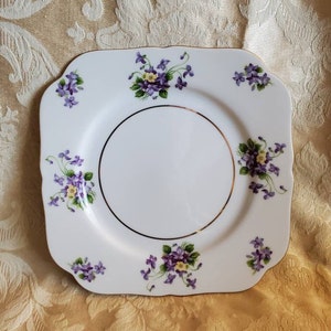 May include: A white square plate with a gold rim and a floral design featuring purple and yellow flowers. The plate has a scalloped edge.