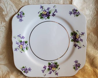May include: A white square plate with a gold rim and a floral design featuring purple and yellow flowers. The plate has a scalloped edge.