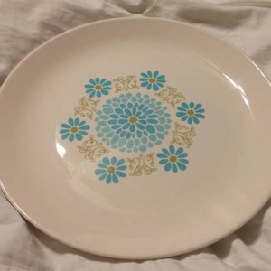 May include: A white ceramic dinner plate with a blue and green floral design. The design features a central flower with smaller flowers surrounding it.
