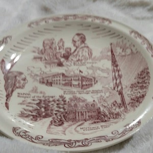 May include: A white oval ceramic plate with a red and white design. The plate features images of historical figures, buildings, and a flag. The text on the plate reads "WADSWORTH, Founder of the Springs, VILLAGE HALL AND MUNICIPAL BUILDING, MY CLARK MANSION, ESTABLISHED 1817".