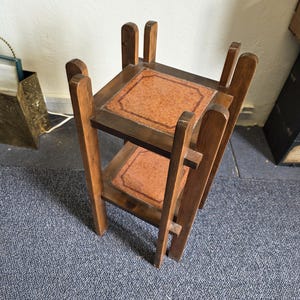 May include: A two-tiered wooden plant stand with a square top and bottom shelf. The shelves have a reddish-brown, patterned inlay. The stand is made of dark wood and has a simple, geometric design. The stand is approximately 24 inches tall.