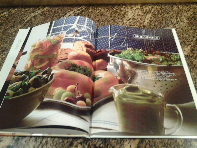 Puede incluir: Un libro de cocina abierto en una p&aacute;gina titulada "Guarniciones". La p&aacute;gina presenta una variedad de alimentos, incluyendo una ensalada en un colador, un taz&oacute;n de jud&iacute;as verdes y una jarra de aderezo. La comida est&aacute; dispuesta sobre un mantel blanco con un fondo de patr&oacute;n azul y blanco.