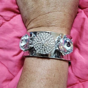 May include: A silver cuff bracelet with three large, clear rhinestone accents. The bracelet is wide and has a smooth, polished finish.