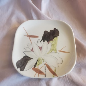 May include: Square ceramic dish with rounded corners. The dish features a white background with a floral design in shades of white, brown, green, and black. The floral design is centered on the dish.