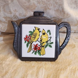 Könnte beinhalten: Gusseisen-Teekannen-Topfuntersetzer mit einer weißen Keramikfliese mit zwei gelben Vögeln mit roten Blumen und grünen Blättern.