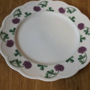 May include: A white ceramic dinner plate with a scalloped edge and a decorative border of green shamrocks and purple flowers.