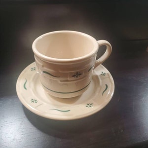 Pode incluir: Uma chávena de chá e um pires de cerâmica branca com um desenho floral verde. A chávena tem uma pega e o pires tem uma borda ondulada.