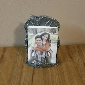 Puede incluir: Un marco de fotos plateado con un diseño de colibrí. El marco contiene una foto de una pareja en un parque.