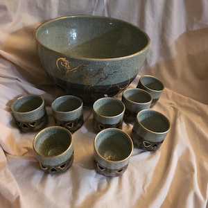 Puede incluir: Un juego de cuenco y tazas a juego de cerámica. El cuenco tiene forma redonda, con un esmalte craquelado de color verde azulado y un diseño de caballo dorado. El juego incluye ocho tazas pequeñas con un diseño similar y ribete dorado.