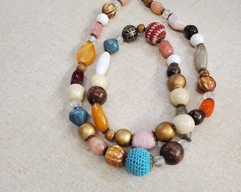 May include: A multi-colored beaded necklace with a variety of textures and shapes, including wood, glass, and metal. The necklace features a mix of colors, including brown, orange, white, blue, and red.