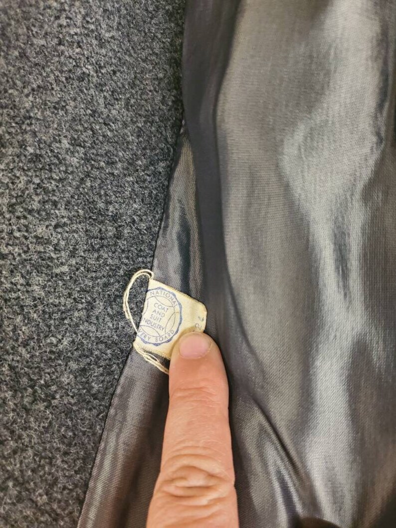 May include: A close-up of a gray wool coat with a label that reads "National Coat and Suit Industry" and "Dry Clean Only".