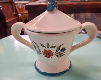 May include: A white ceramic sugar bowl with a blue and floral design. The bowl has two handles and a lid with a blue knob.