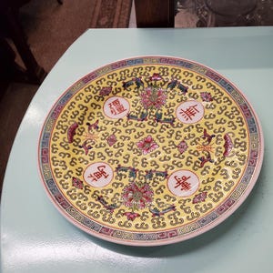May include: A yellow ceramic plate with a decorative border and floral design. The plate features four red Chinese characters in circles.