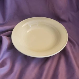 Op de afbeelding: Een eenvoudige witte keramische kom met een licht verhoogde rand. De kom staat op een paarse stoffen ondergrond.