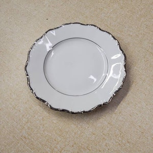 Könnte beinhalten: Ein weißer Porzellanteller mit silbernem Rand und einem dünnen silbernen Band nahe der Mitte. Der Teller hat einen gewellten Rand und ist vor einem neutralen Hintergrund platziert. Dieser elegante Teller eignet sich zum Servieren von Vorspeisen oder Desserts.