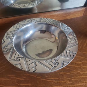 Könnte beinhalten: Eine silberne Schale mit einem geometrischen Muster am Rand. Die Schale ist aus Metall und hat eine glänzende Oberfläche.