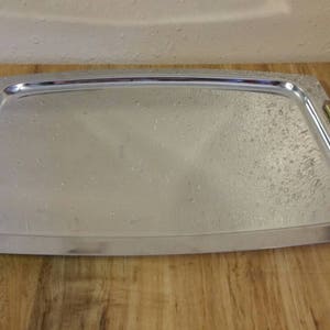 May include: A rectangular silver serving tray with wooden and brass handles. The tray has a slightly textured surface and a raised edge. The handles are rectangular with a brass accent. The tray is on a wooden surface.