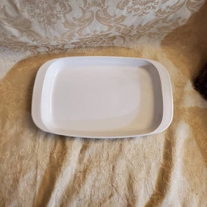 Puede incluir: Plato rectangular para hornear blanco con un borde elevado. El plato está hecho de cerámica y tiene un acabado liso y brillante.