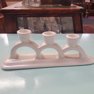 May include: White ceramic candle holder with three candle cups. The holder has a curved design with three arches.