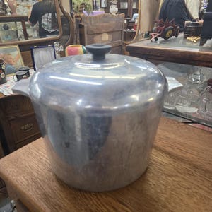 May include: A silver-colored aluminum pot with a matching lid and a black knob handle on top. The pot features two side handles and has a tapered, cylindrical body with a smooth, metallic finish, showing signs of light wear and surface texture.
