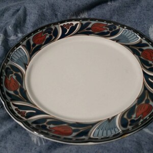 May include: A white ceramic dinner plate with a blue and red floral design. The plate has a scalloped edge.