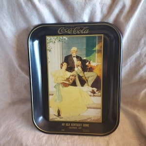 May include: A black metal tray with a Coca-Cola logo and a vintage illustration of a couple sitting on a porch. The illustration is titled "My Old Kentucky Home 1931 Calendar Art".