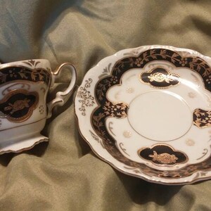 May include: A white teacup and saucer set with black and gold floral designs. The cup has a handle and the saucer has a scalloped edge.