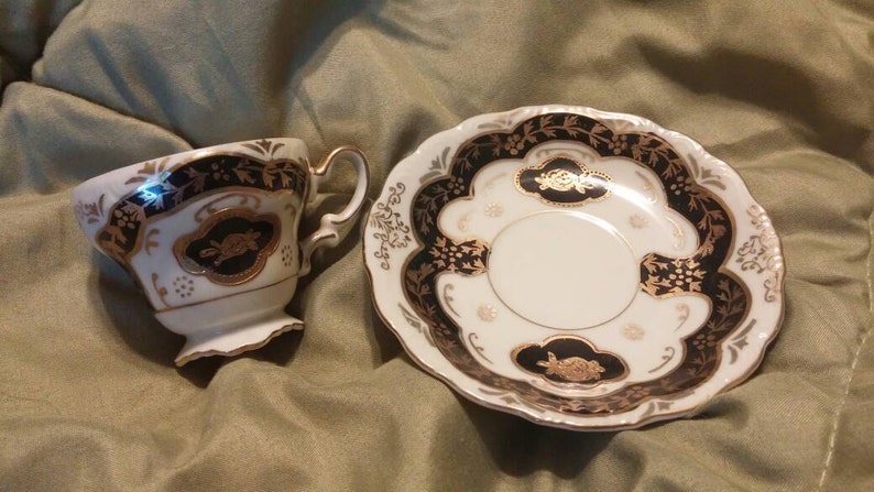 May include: A white teacup and saucer set with black and gold floral designs. The cup has a handle and the saucer has a scalloped edge.