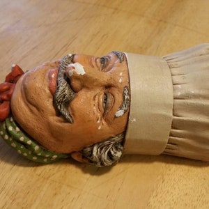 May include: A ceramic figurine of a chef's head wearing a white toque and a red and green patterned bandana. The chef has a moustache and a friendly expression.