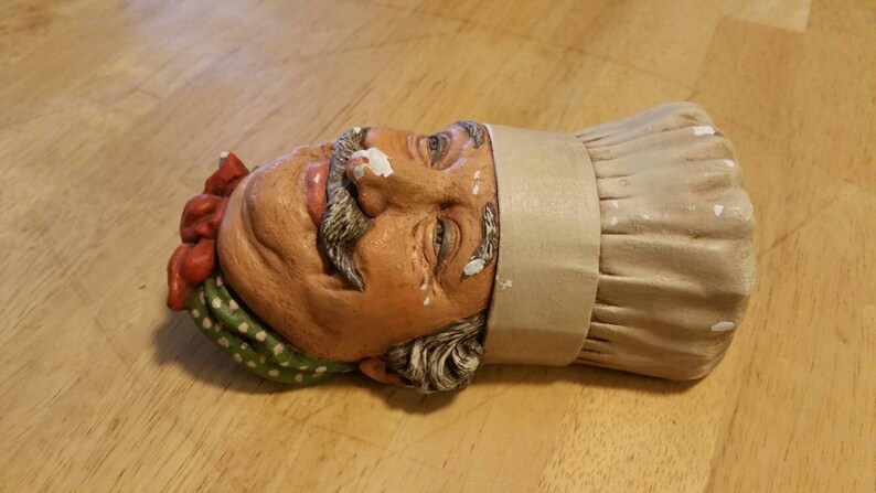 May include: A ceramic figurine of a chef's head wearing a white toque and a red and green patterned bandana. The chef has a moustache and a friendly expression.