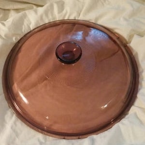 Op de afbeelding: Een doorzichtig glazen deksel met een paarse rand en knop. Het deksel heeft een getextureerd oppervlak en is ontworpen voor een ovenschaal.